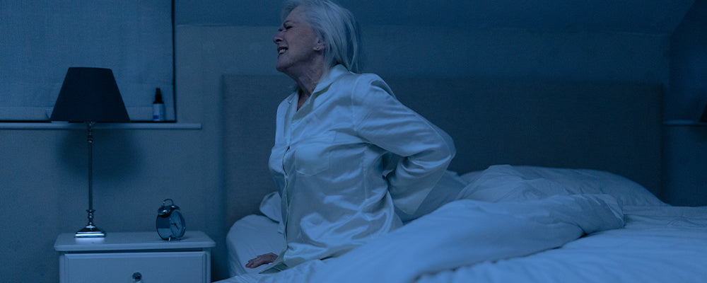 Older woman sat at the edge of bed in pain with one arm holding her back