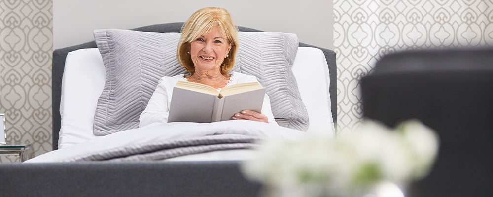 Elderly woman in bed smiling with a book in her hand and the back of the bed is rasing so she is sitting up