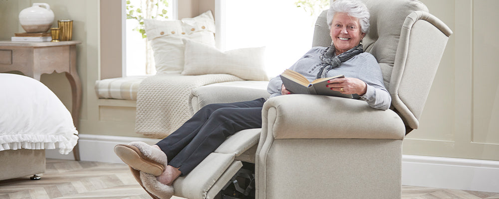 older woman sat in an Opera riser recliner chair with her legs up smiling holding a book