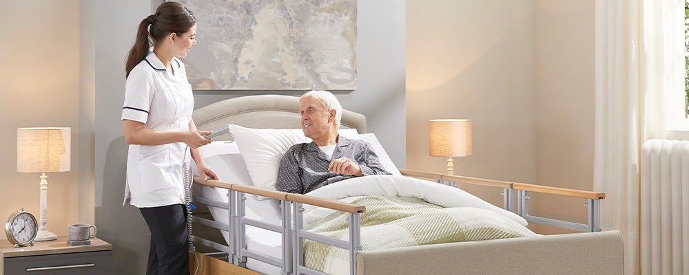 Younger woman in a carers uniform stood holding a handset next to a care be. In the care bed is a older gentleman who is looking at the carer