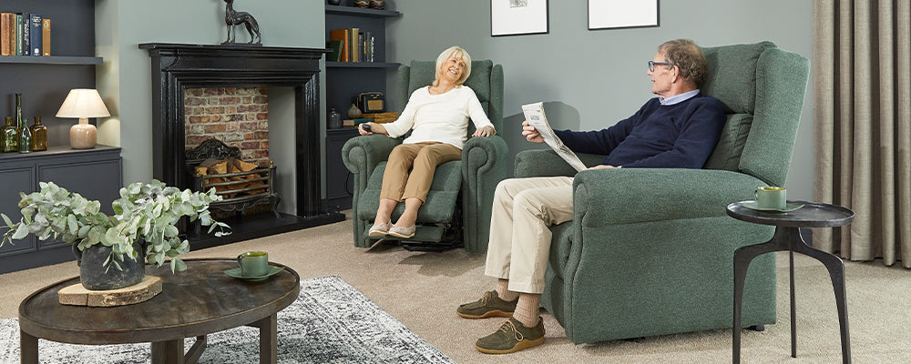 A older woman and gentleman sat in a living room in a riser recliner chair, the lads is sat reclined with her legs up and the gentleman is sat up reading a newspaper