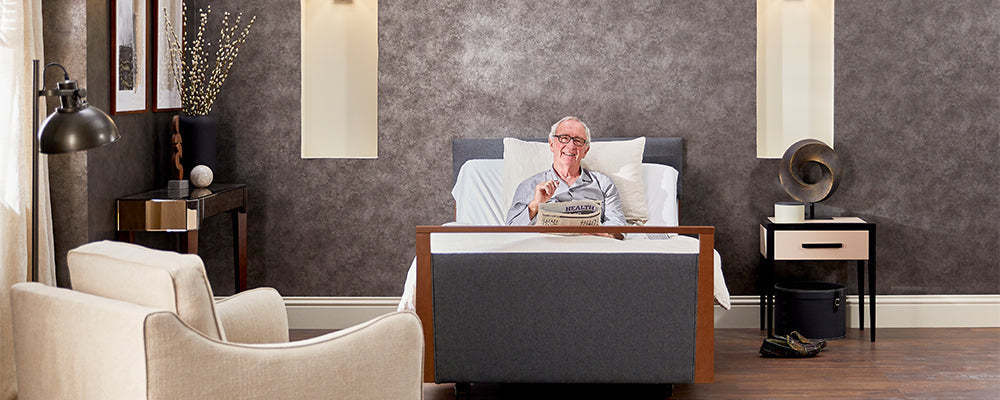 A front view of a older gentleman sat upright in a profiling bed smiling and holding a newspaper