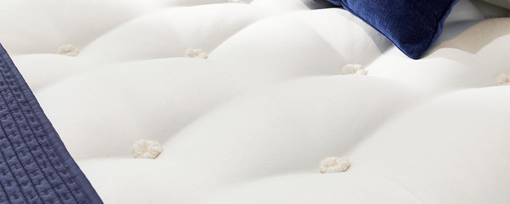 Close up of a white quilted mattress with a navy blue scatter cushion and blue throw on the bottom of the mattress