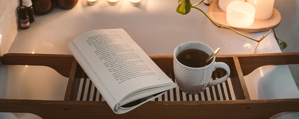 Bath tray with coffee cup and open book with lit pillar candles in the background