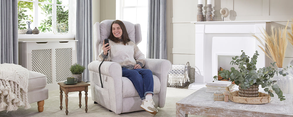 A woman sat in a riser recliner chair smiling and holding up the wired hand control