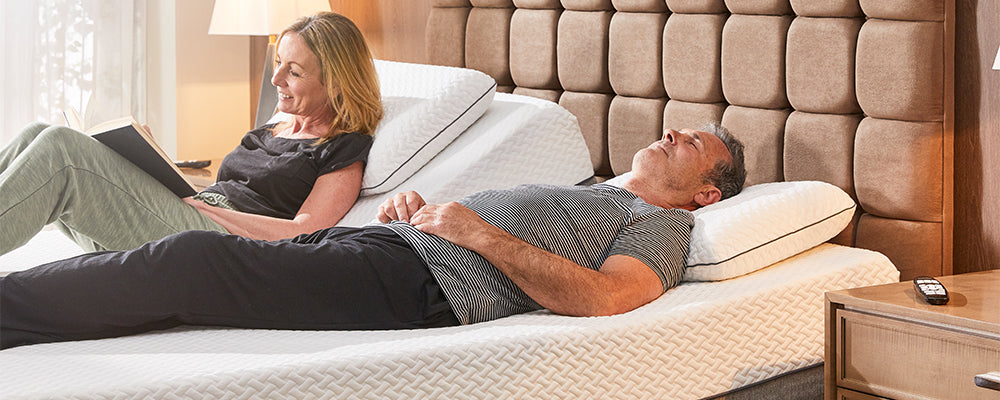 man and woman laid in an adjustable bed, the woman is reading a book whilst the gentleman is asleep with his head rest section raised slightly