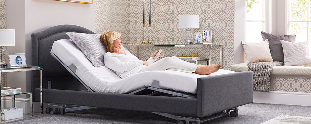 Lady with blonde hair sat upright in a profiling bed with her legs raised and holdimg the remote control