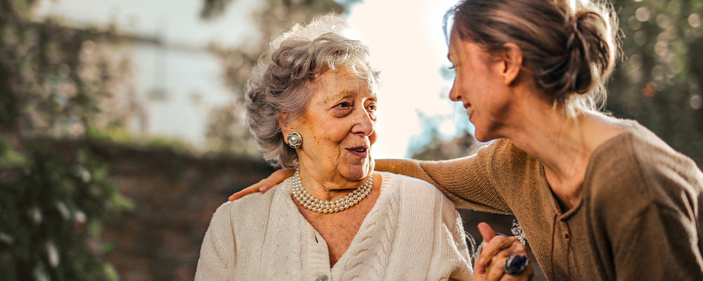 Older lady wearing sparkly necklace and earrings sat with a younger female with her arm around her