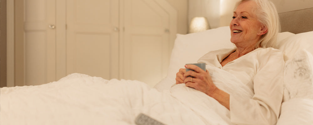 Elderly woman sat upright in bed smiling and holding a blue mug