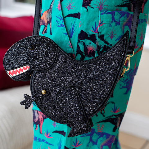 The kawaii black glitter dino vegan bag being worn by a model wearing dino print dungarees. The bag is facing forward to highlight the dinosaur face, moveable arm, detachable strap and black glitter front.