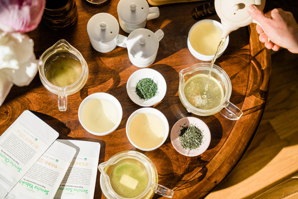 An array of tea pots and tea leaves cover a wooden table where sencha Japanese tea is being brewed