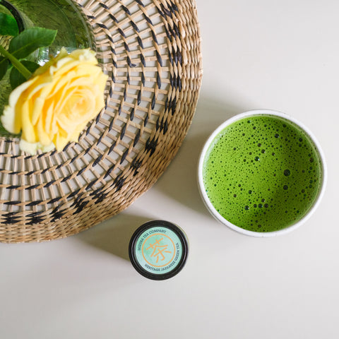 An overhead view of a bowl of matcha tea next to a woven placemat where a yellow rose in a vase sits