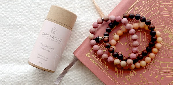 Compostable deodorant tube with pink label laying next to pink notebook and 2 beaded bracelets