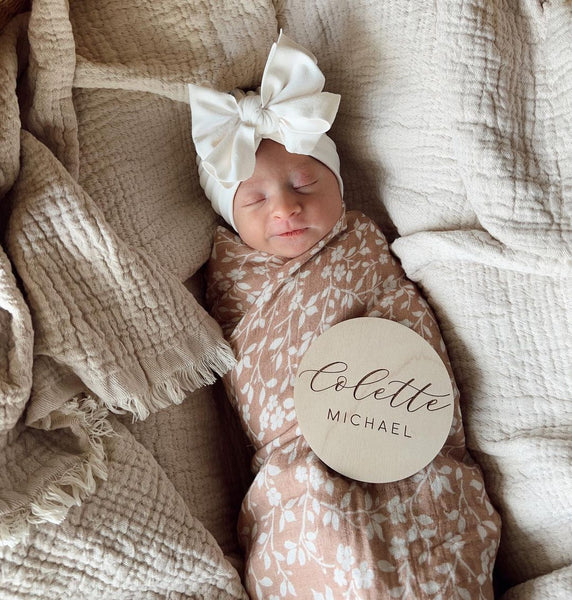 Baby girl smiling while asleep swaddled in mini wander Magnolia Tree Brown, wearing a big white bow.  She's dozing off on a soft brown cotton linen.