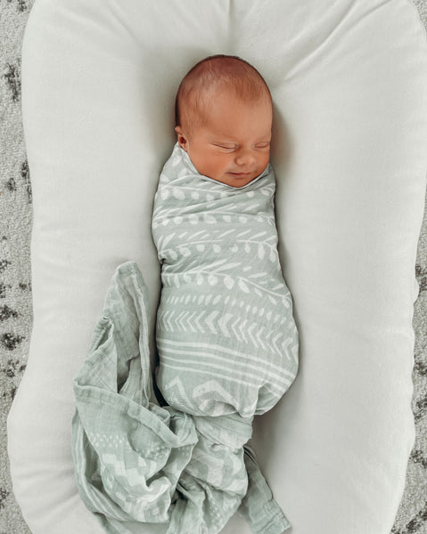 a smiling newborn baby boy swaddled in a green boho wrap in a snuggle me organic bassinet. 