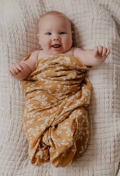 smiling baby, wrapped in a mustard color swaddle with leaf design, the baby was placed in a baby lounger covered with knitted blanket