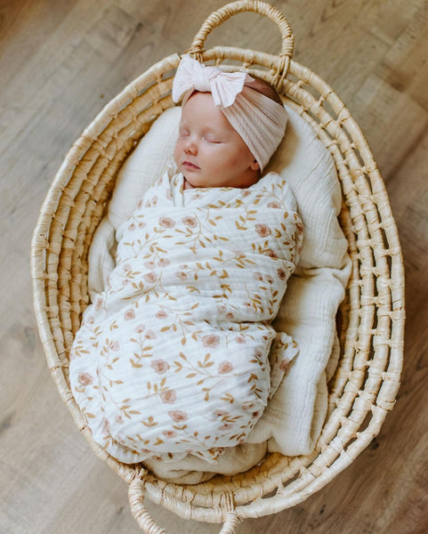 Baby sleeps soundly in a baby basket while swaddled in a mini wander wildflower white swaddle with a big light peach bow.