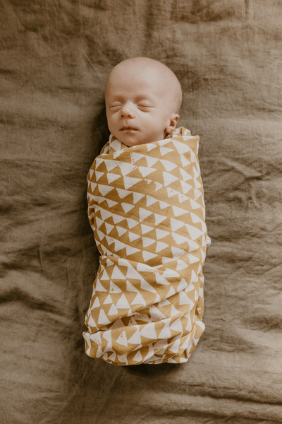 newborn baby boy swaddled in a dark yellow triangle, swaddle with a mountain pattern, taking a baby nap on a bed covered with solid color brown bed sheet