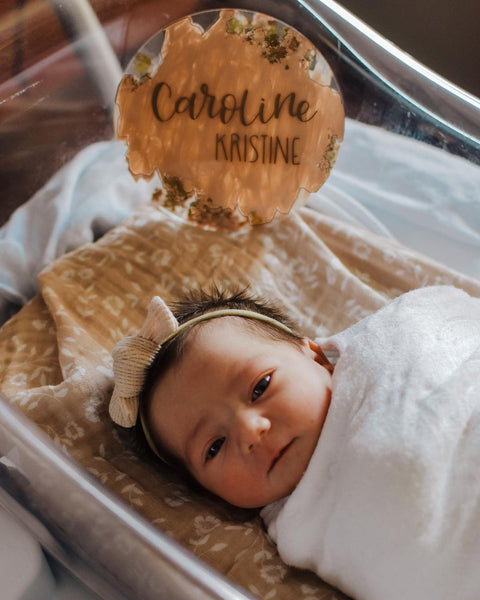 Newborn baby girl named Caroline Kristine, swaddled in white swaddle sleeping in a Hospital bassinet covered with mini wander wild flower taupe swaddle. The baby has a lovely light brown hair bow.