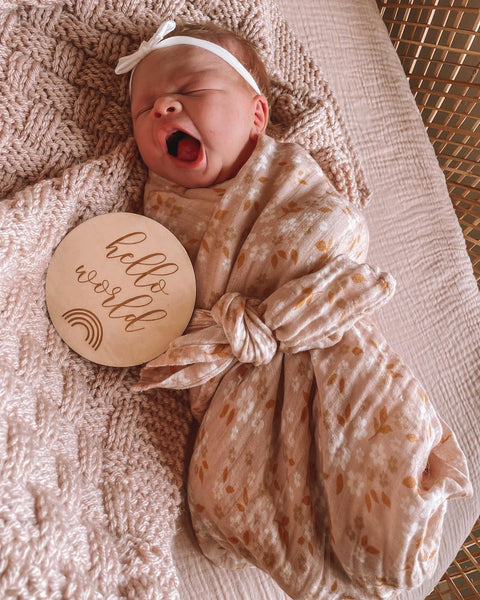 Wayning, a newborn baby girl wrapped in a pink flower swaddle, with a hello world wooden board announcement next to her. She's got a cute white bow on her head.