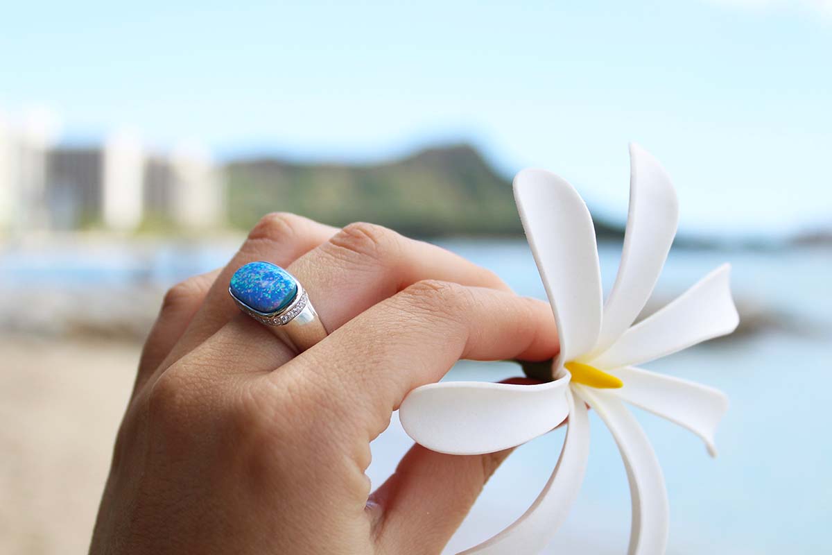 A hand wearing an opalite ring, holding a white flower with a beach background.