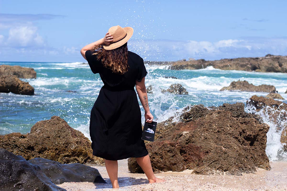 Helen Stephenson facing the ocean and holding her book when the tide rolls in while wearing a larimar ring on a beach