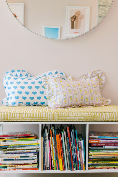 white metal bench with stacks of books and a yellow upholstered cushion on top with two toddler sized cotton ruffle pillows on top of the cushion. one pillow is white with blue hearts. the other pillow is pink with a small floral design. 