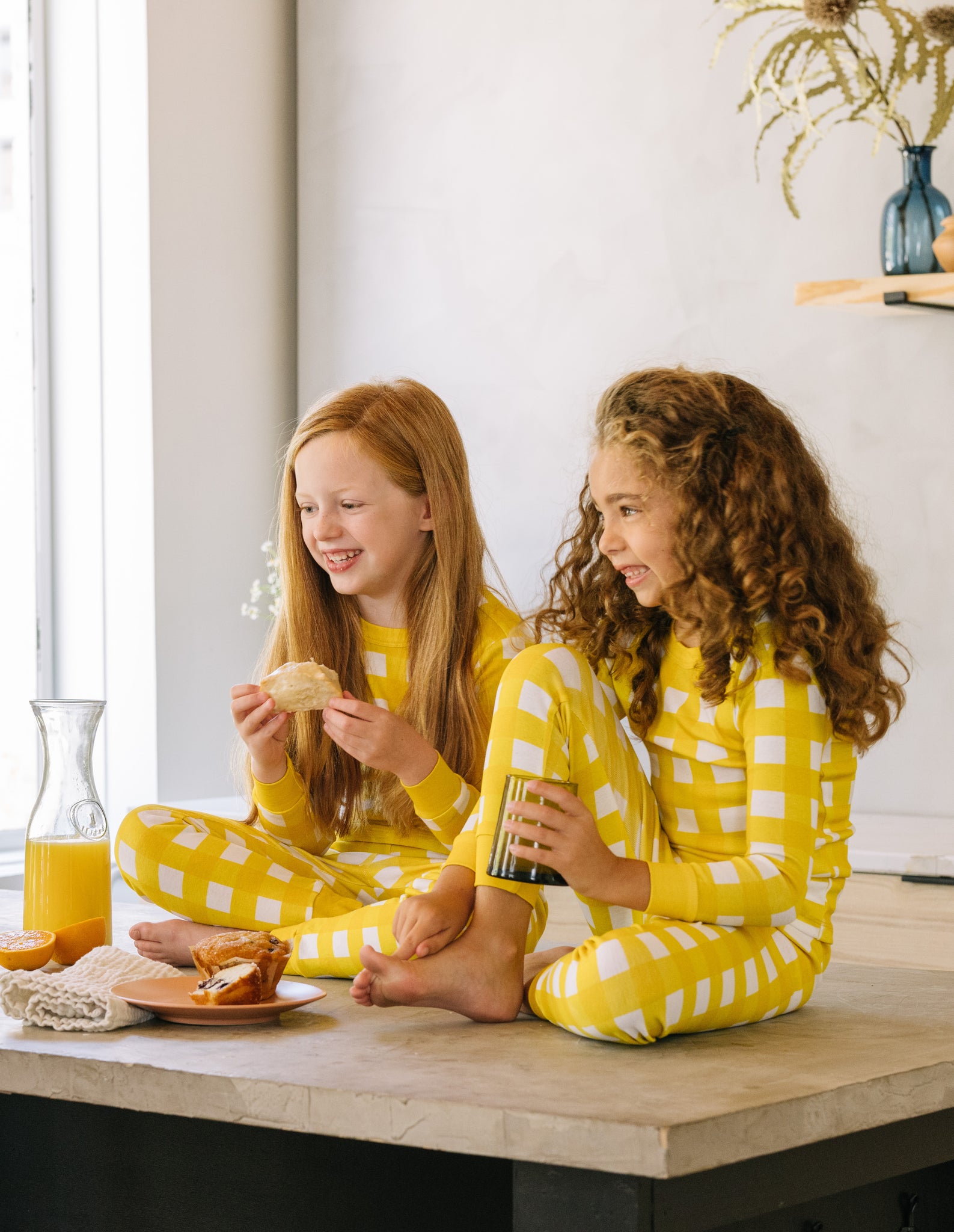 soft, yellow gingham pajama sets worn by two girls drinking orange juice and eating breakfast