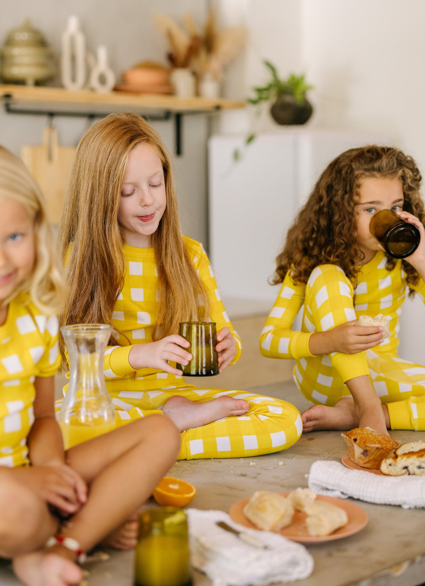 soft, yellow gingham kids pajama sets worn by three girls drinking orange juice and eating breakfast