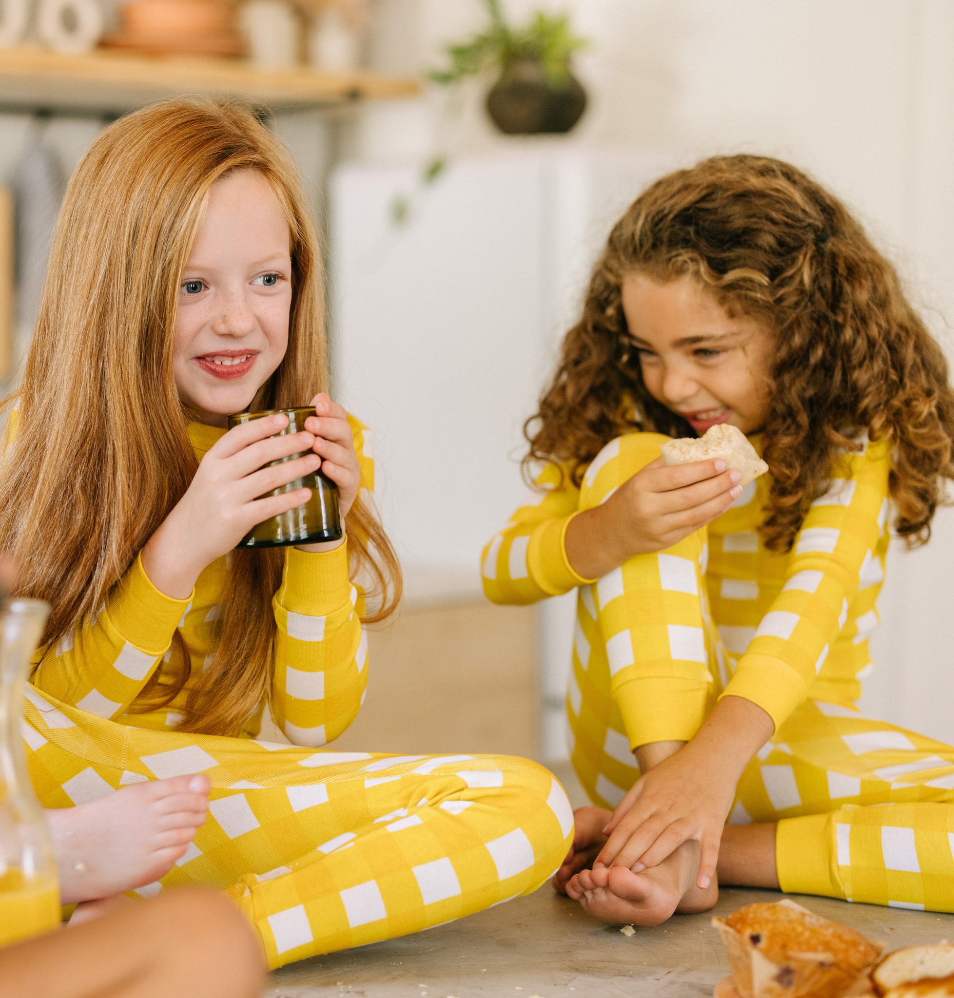 soft, yellow gingham pajama sets worn by two girls drinking orange juice and eating breakfast