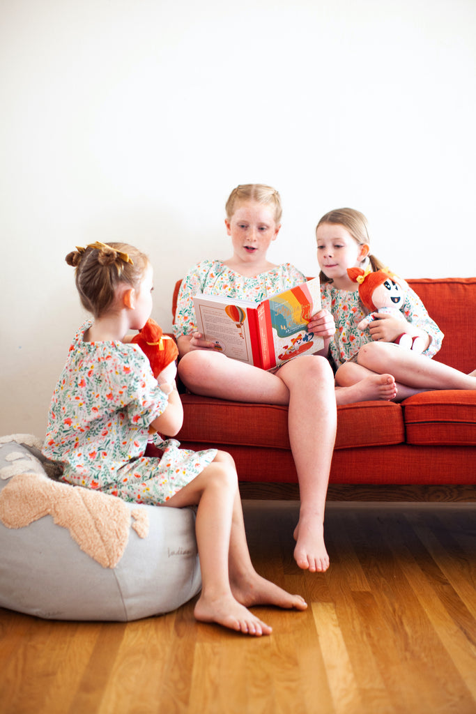 big sister reading to little sister. all wearing matching floral cotton dresses.