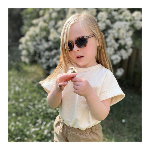 Petite fille avec lunette de soleil qui tient une fleurs dans les mains et habillée avec le t-shirt Antero de chez Mom(e) Portugal ainsi que du pantalon en lin de chez Omibia