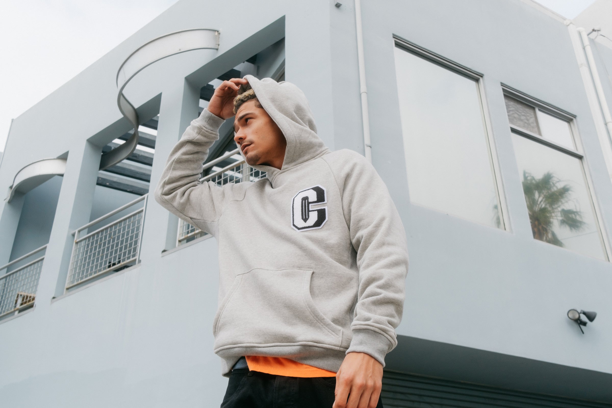 Model wearing the Varsity C Hoodie in grey and standing in front of a building.