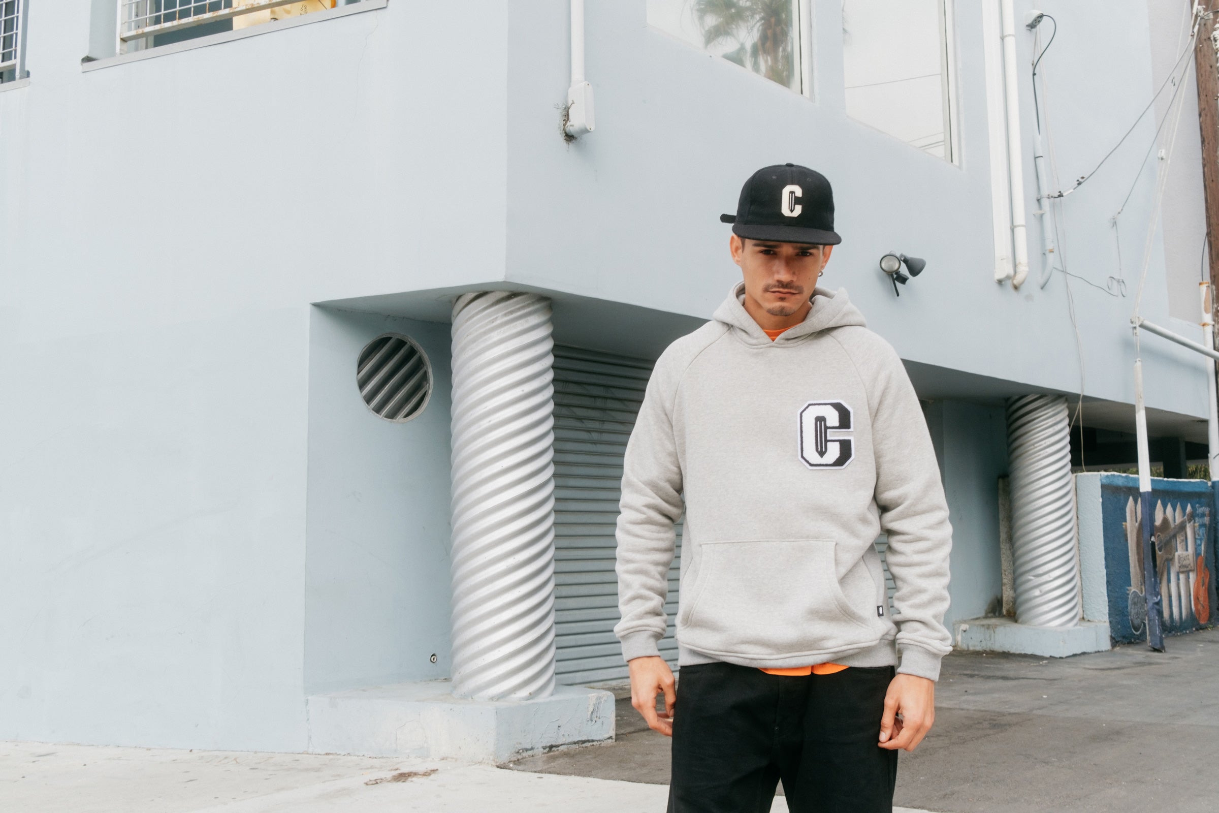 Model wearing the Varsity C Hoodie in grey and the Ebbets Wool C Cap in black and standing in front of a building.