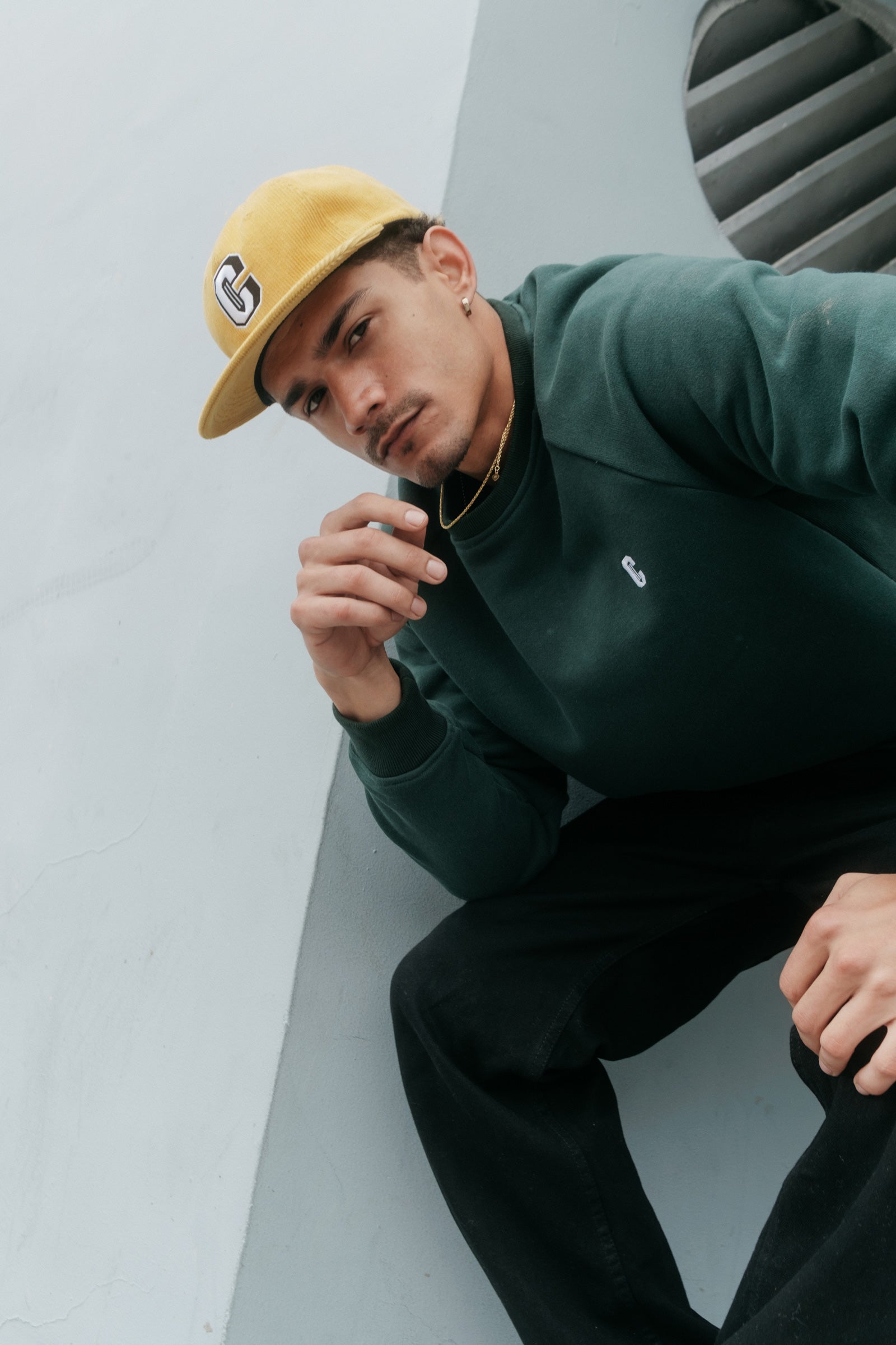 Model wearing the Varsity C Corduroy Cap in yellow and Icon Crewneck Sweatshirt in forest green and sitting at the corner of a building.