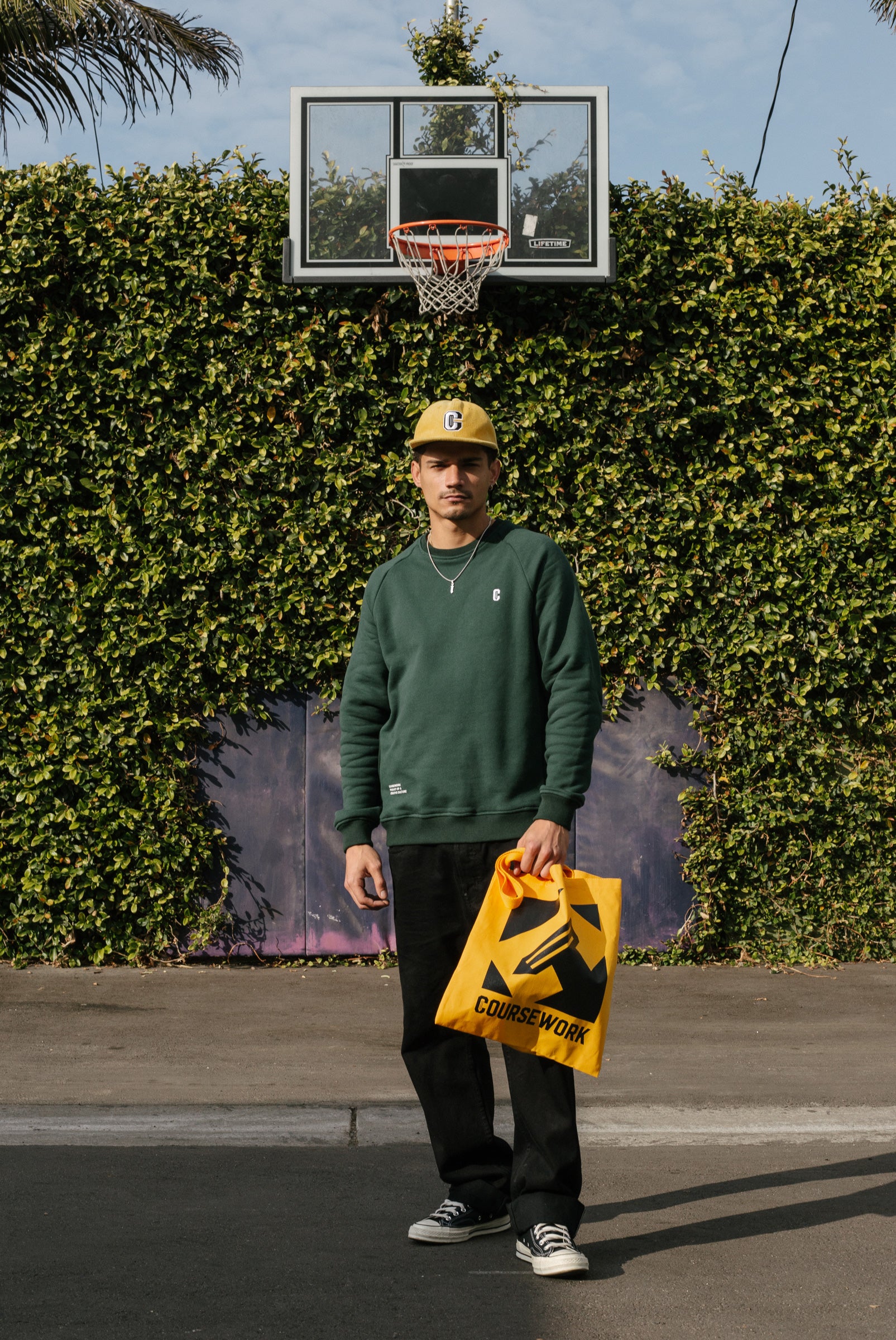 Model wearing the Varsity C Corduroy Cap in yellow and Icon Crewneck Sweatshirt in forest green and standing in front a basketball hoop and foliage..