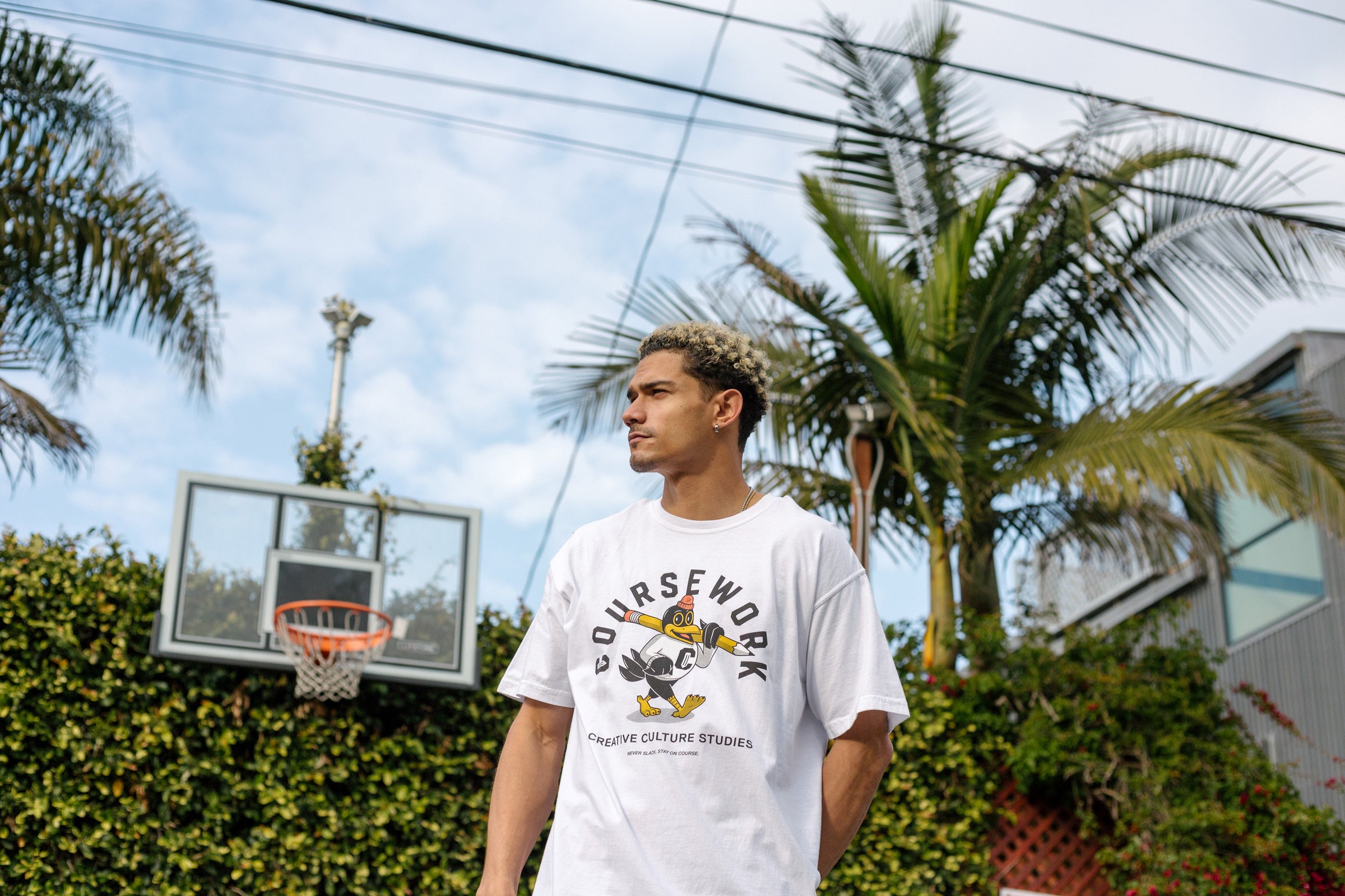Model wearing the Early Birds tee with a basketball hoop, palm tree and green foliage behind him.
