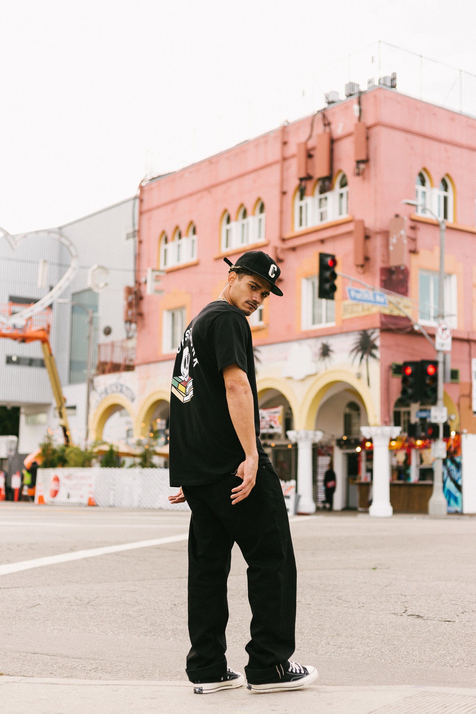 Model wearing the Ebbets Wool C Cap in black and the Required Reading Tee in black while standing on the sidewalk of an intersection.