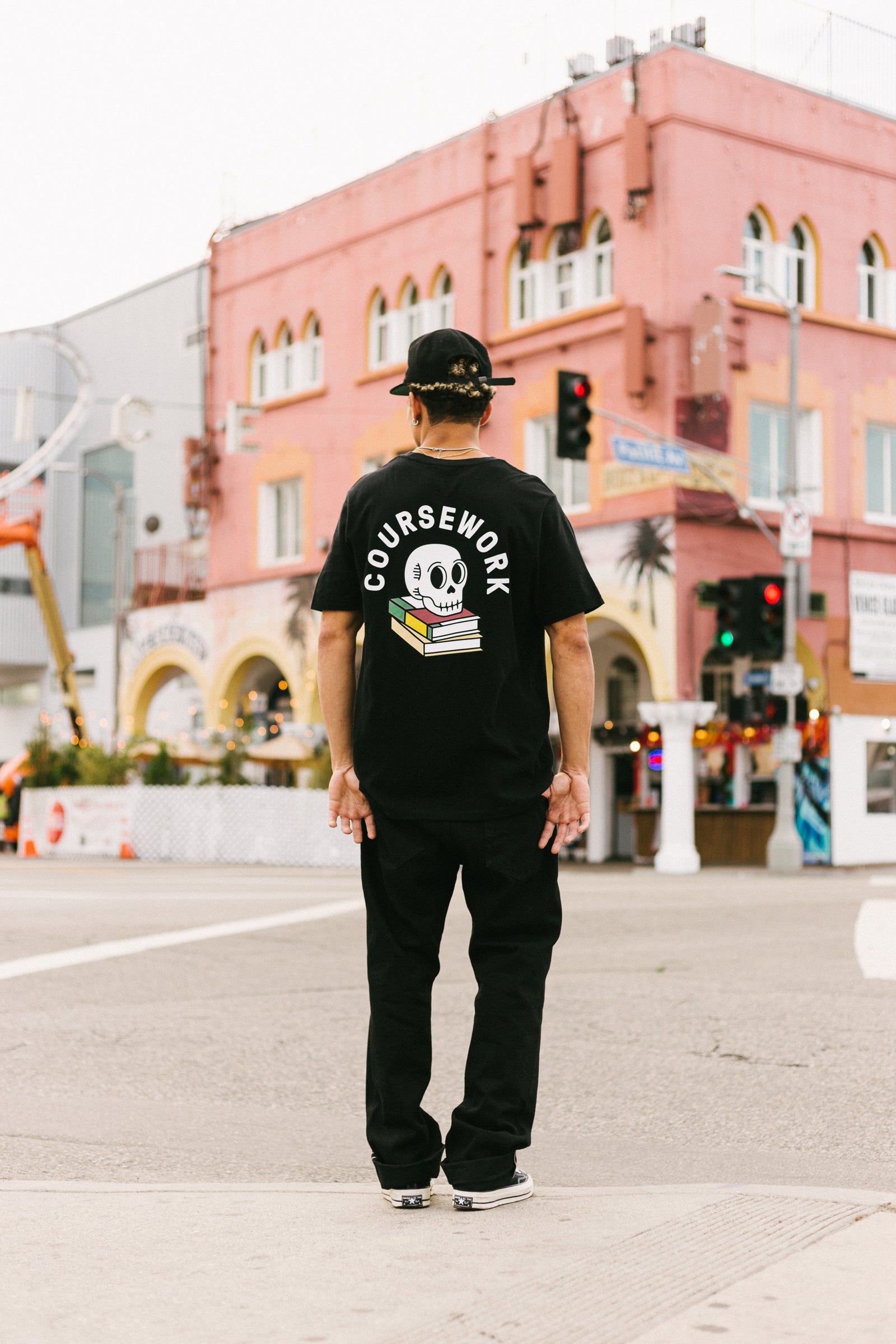 Model wearing the Ebbets Wool C Cap in black and the Required Reading Tee in black while standing on the sidewalk of an intersection.