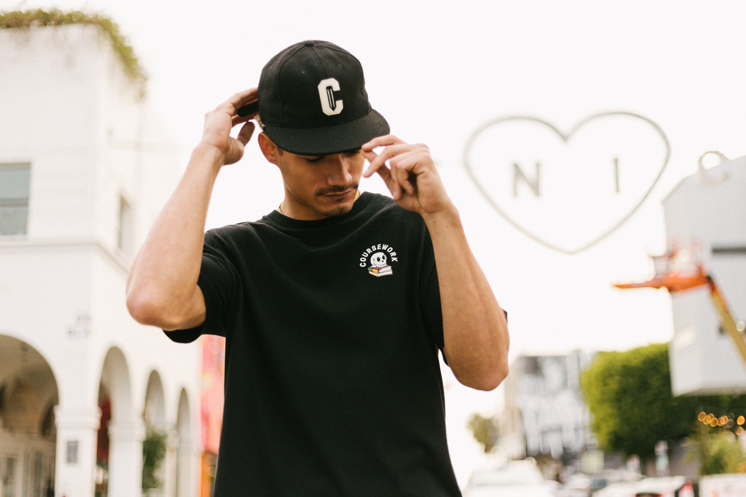 Model wearing the Ebbets Wool C Cap in black and the Required Reading Tee in black while standing on the sidewalk of an intersection.