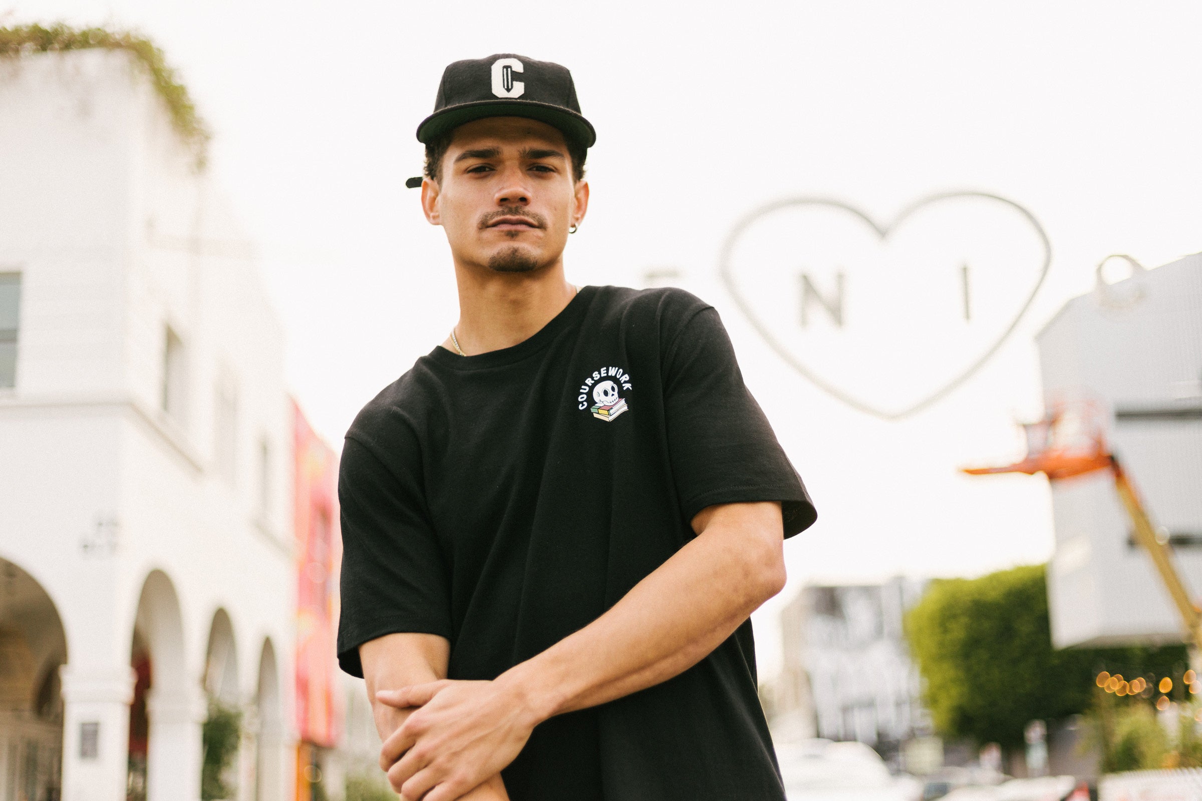 Model wearing the Ebbets Wool C Cap in black and the Required Reading Tee in black while standing on the sidewalk of an intersection.