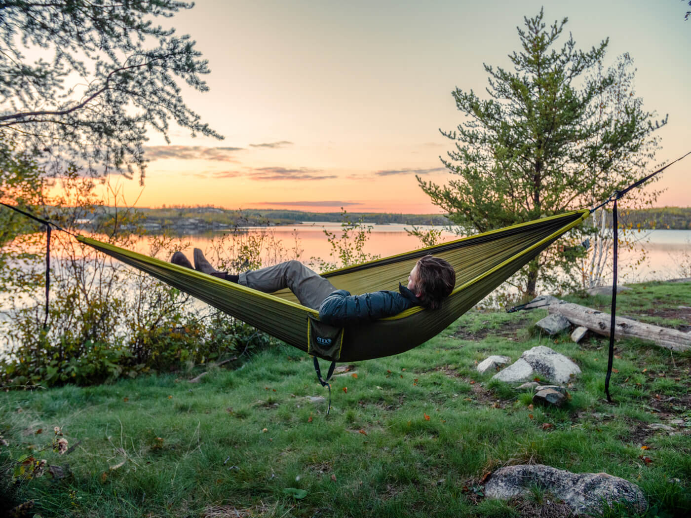 A man lays in a hammock near a lake at sunset. 