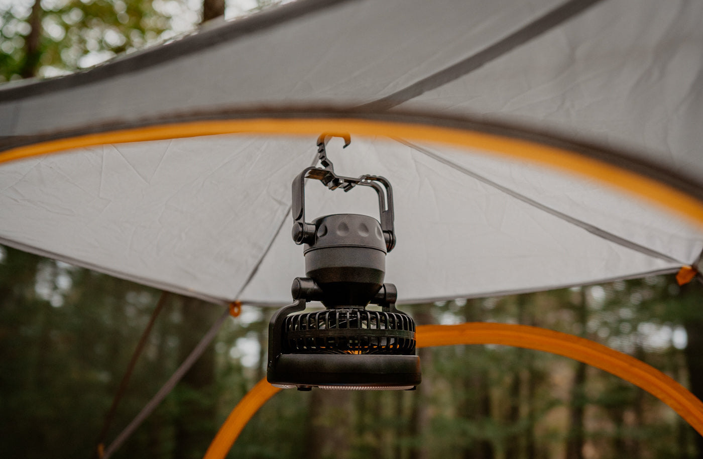 A portable fan hangs from the roof of a tent at a campsite. 