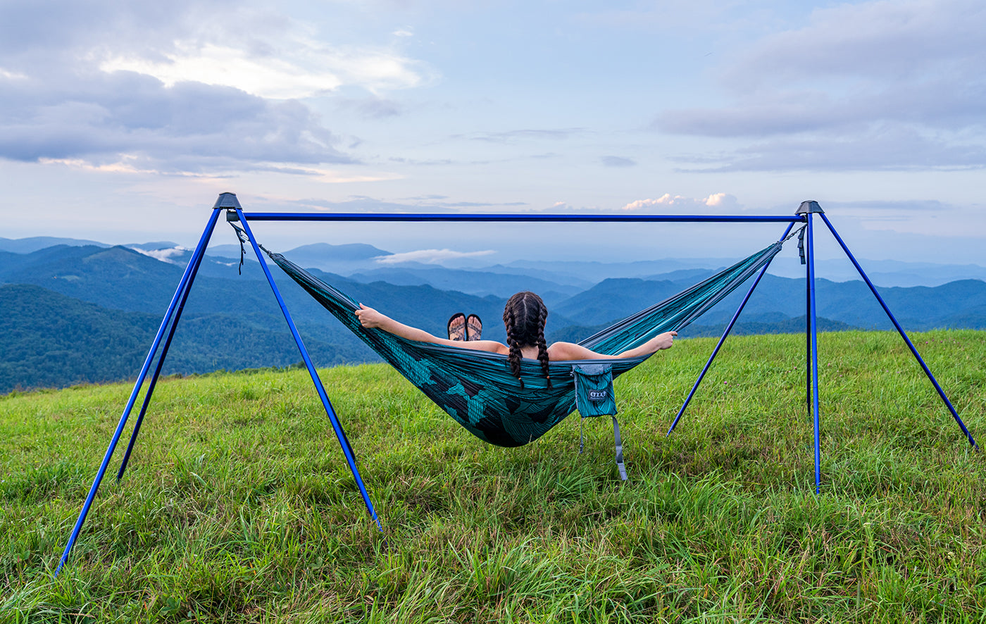 A woman sits in a DoubleNest Hammock Print while using the Nomad Hammock Stand overlooking the Blue Ridge Mountains.