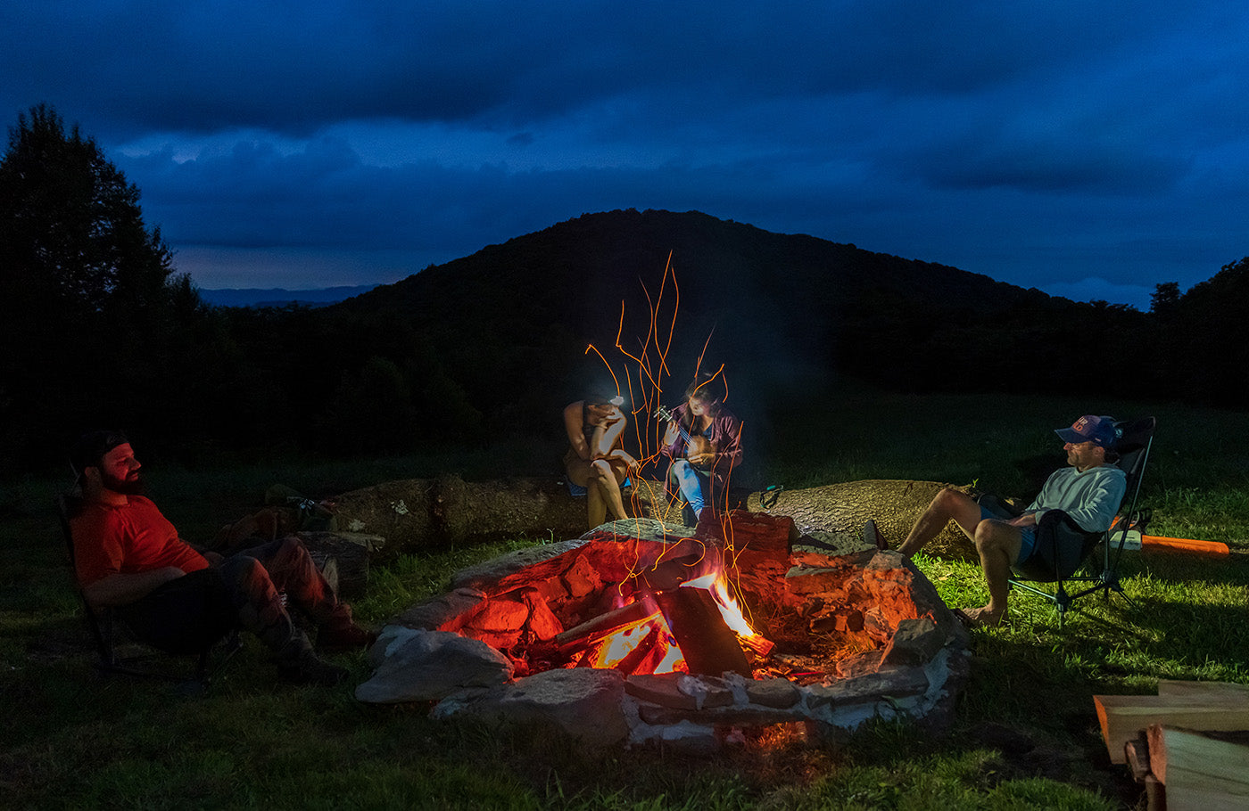 Three people sit around a campfire at dusk. 