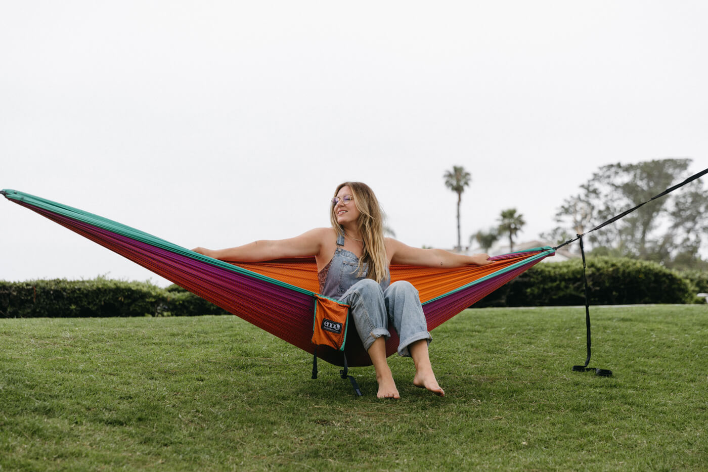 A woman sits in her Fade DoubleNest Hammock Print near the coast.