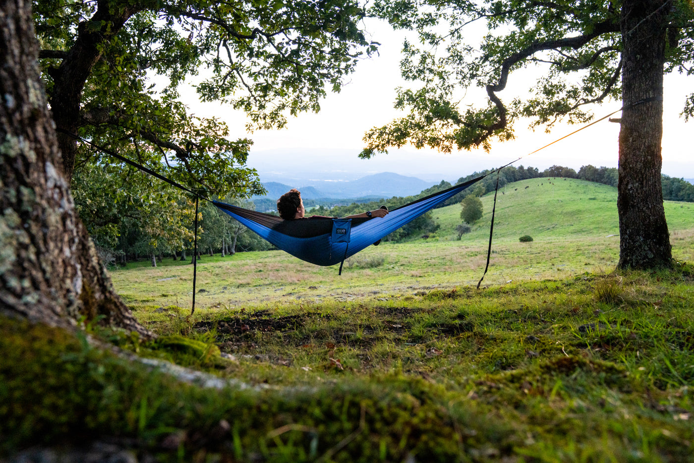 A man sits in a blue DoubleNest hammock that overlooks the blue ridge mountains.