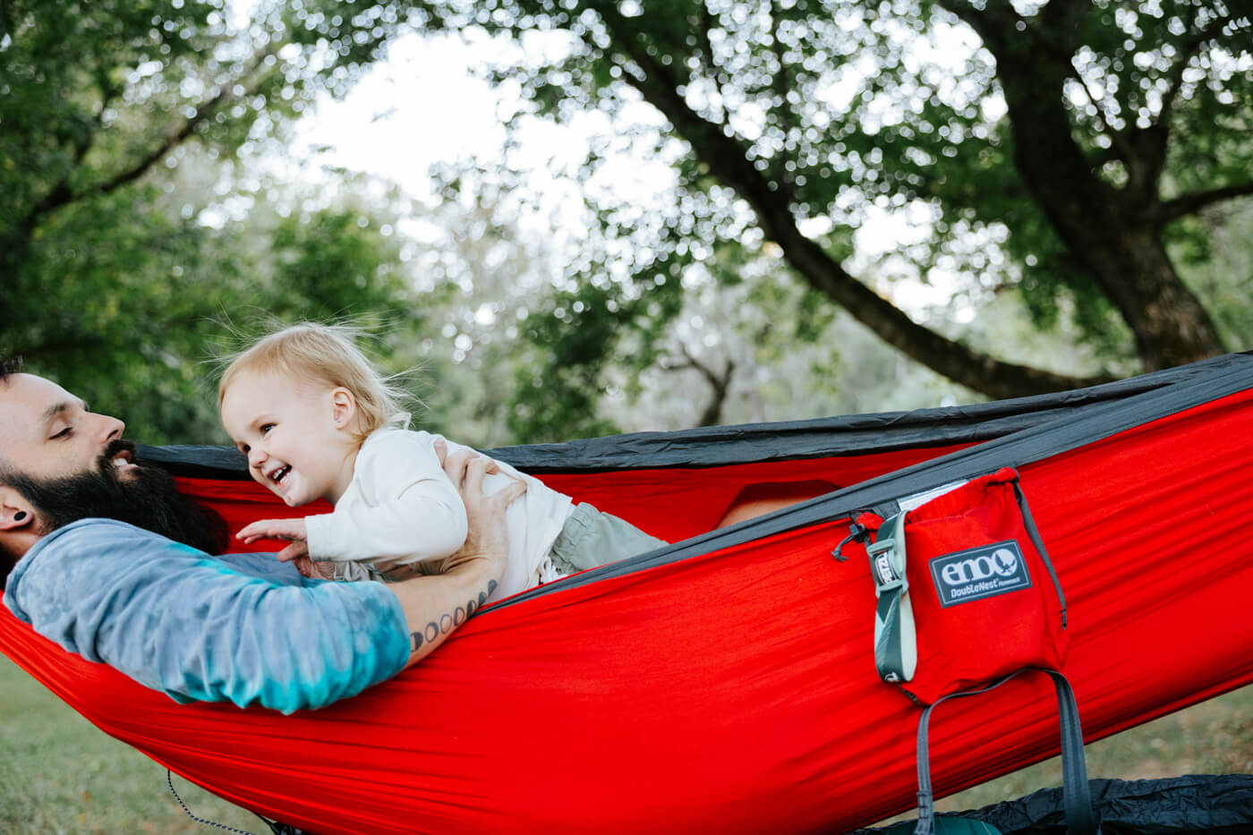 A man plays with his kid while laying in a red ENO hammock together