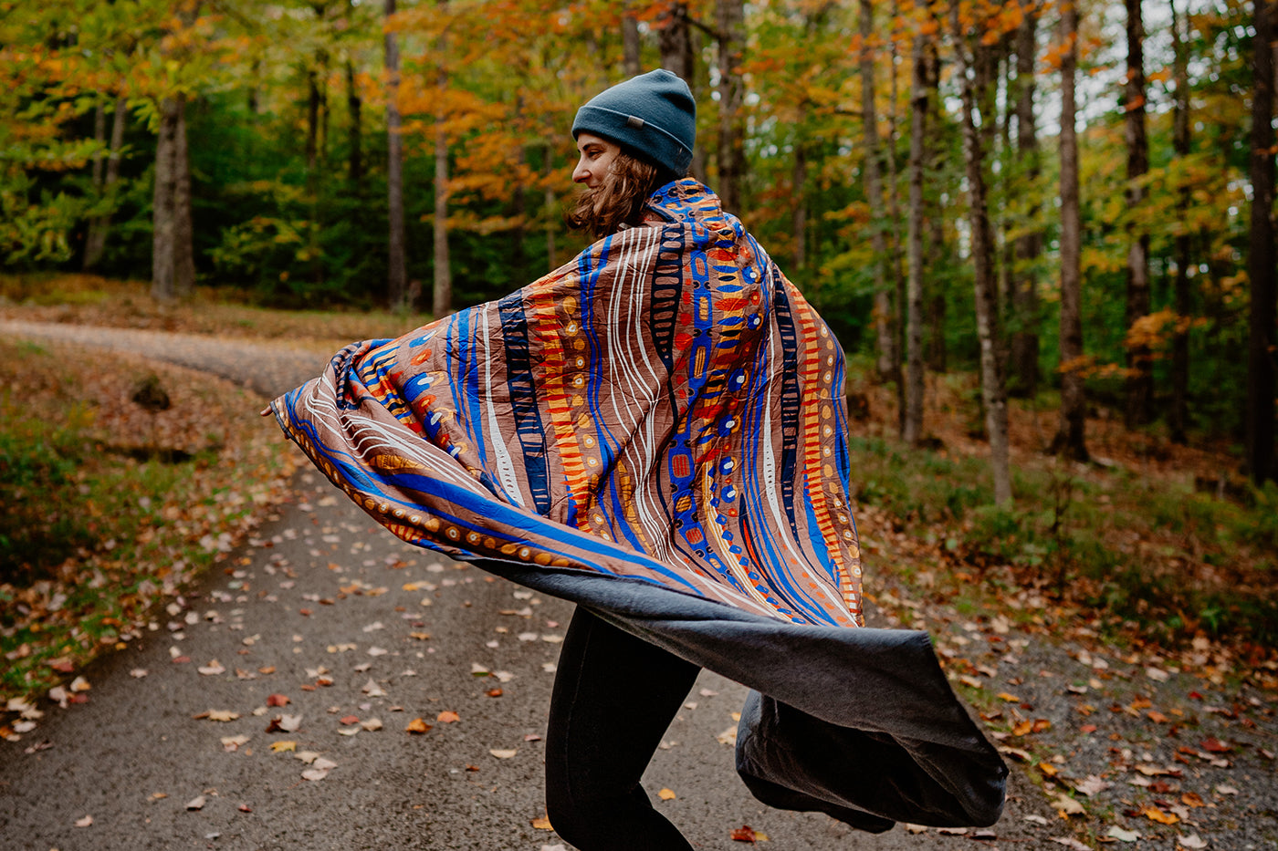 A woman spins while wearing the FieldDay Blanket surrounded by fall foliage.