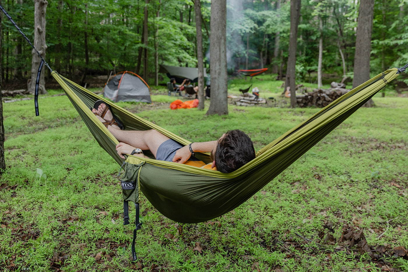 A man sits in a DoubleNest hammock while camping